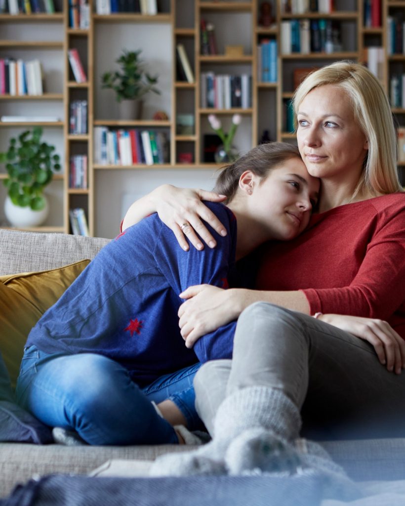 Mother and adolescent daughter sitting on couch with arms around